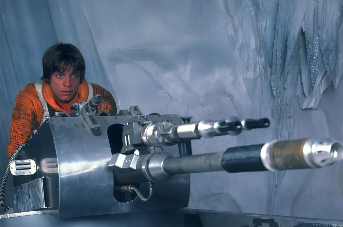A young man in an orange flight suit peers nervously from behind a large, complex, metallic weapon mounted on a vehicle, set…