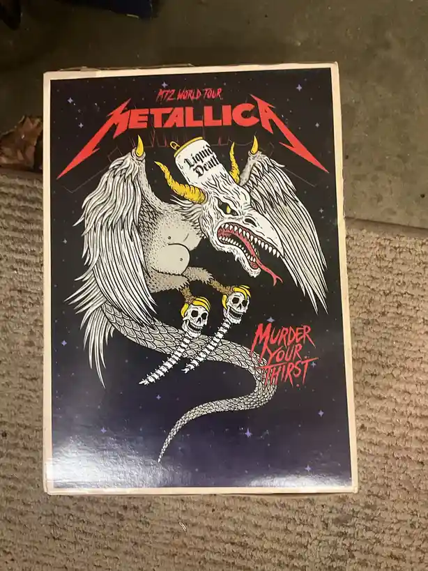 A cardboard box featuring a Metallica 'MTZ World Tour' poster with a demonic, winged creature holding a can labeled 'Liquid…