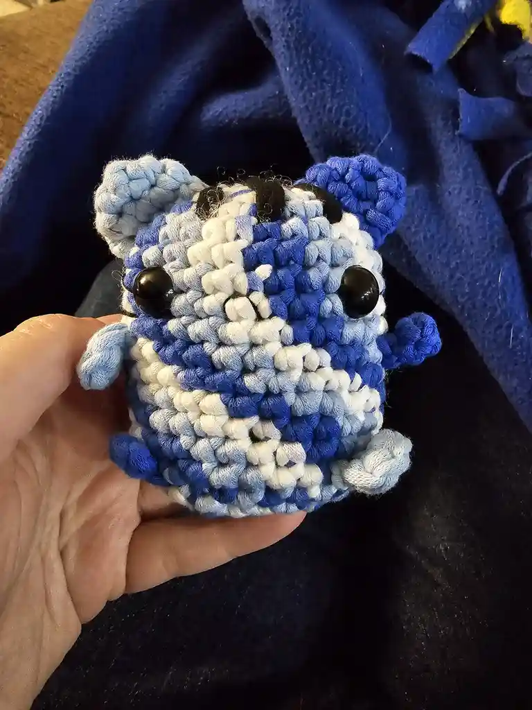 A hand holds a small, crocheted plush toy made of blue, white, and light blue yarn, featuring large black button eyes and a…
