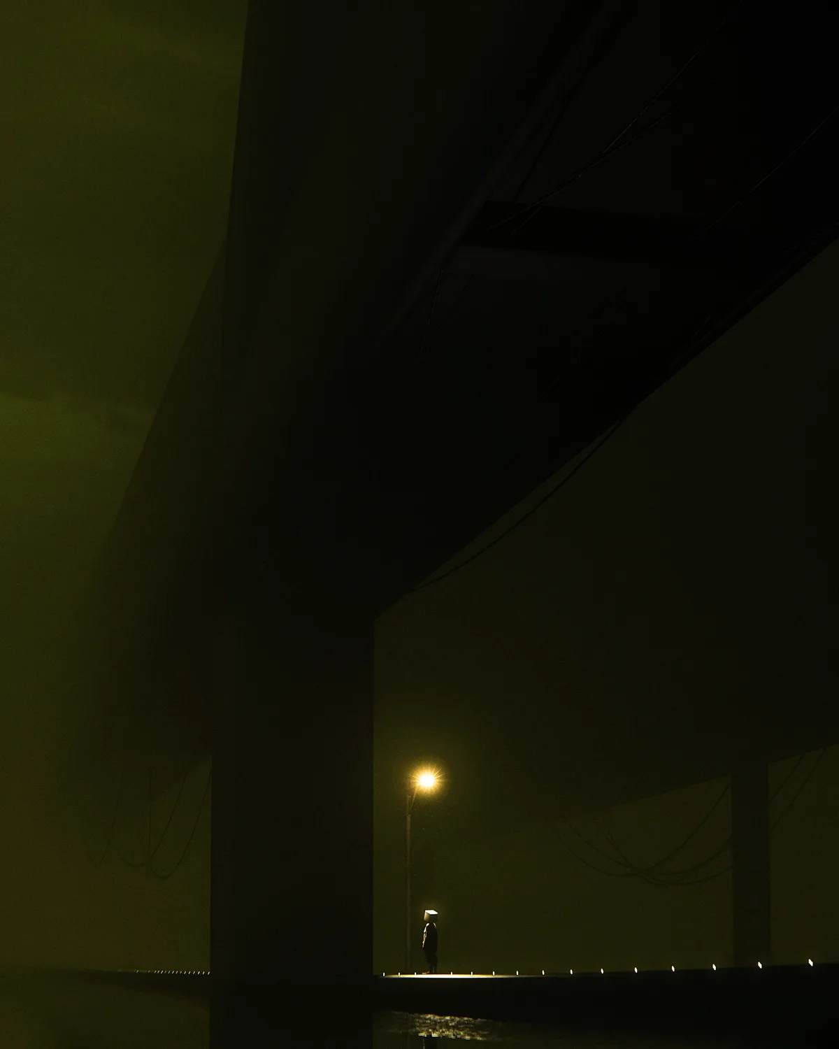A solitary figure stands under a dim, yellow streetlight beneath a large overpass at night, their head covered by a white…