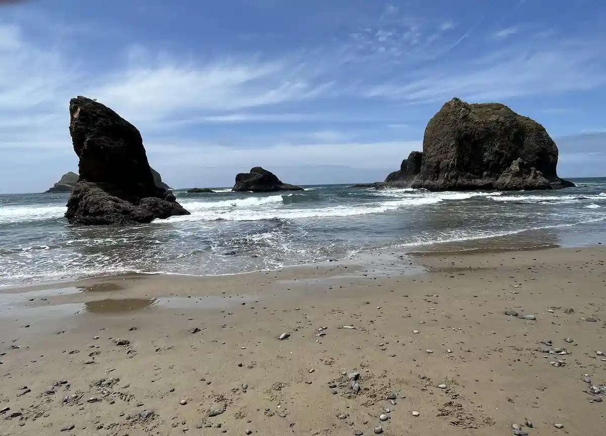 A scenic beach photo shows waves crashing against large, dark sea stacks under a partly cloudy blue sky. The foreground is…