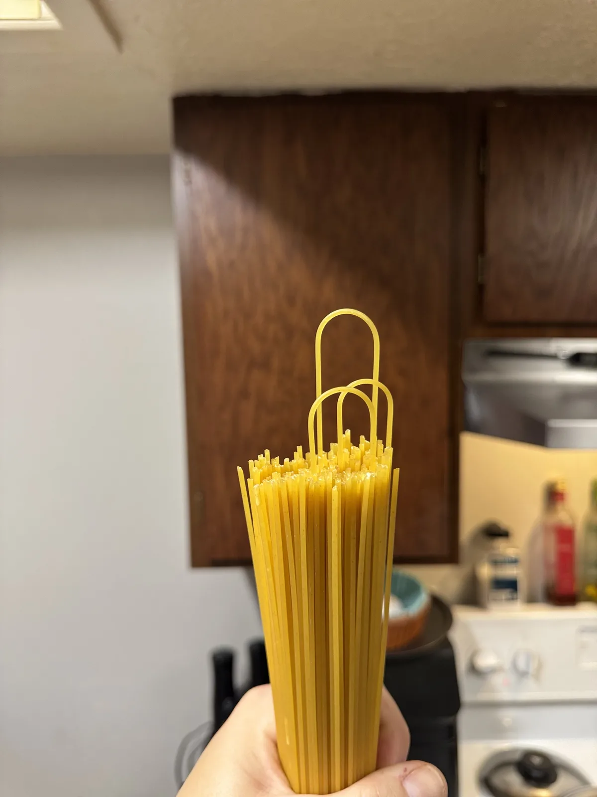 A hand holds a bundle of uncooked spaghetti in a kitchen, with the top two strands bent into the shape of paperclip loops…