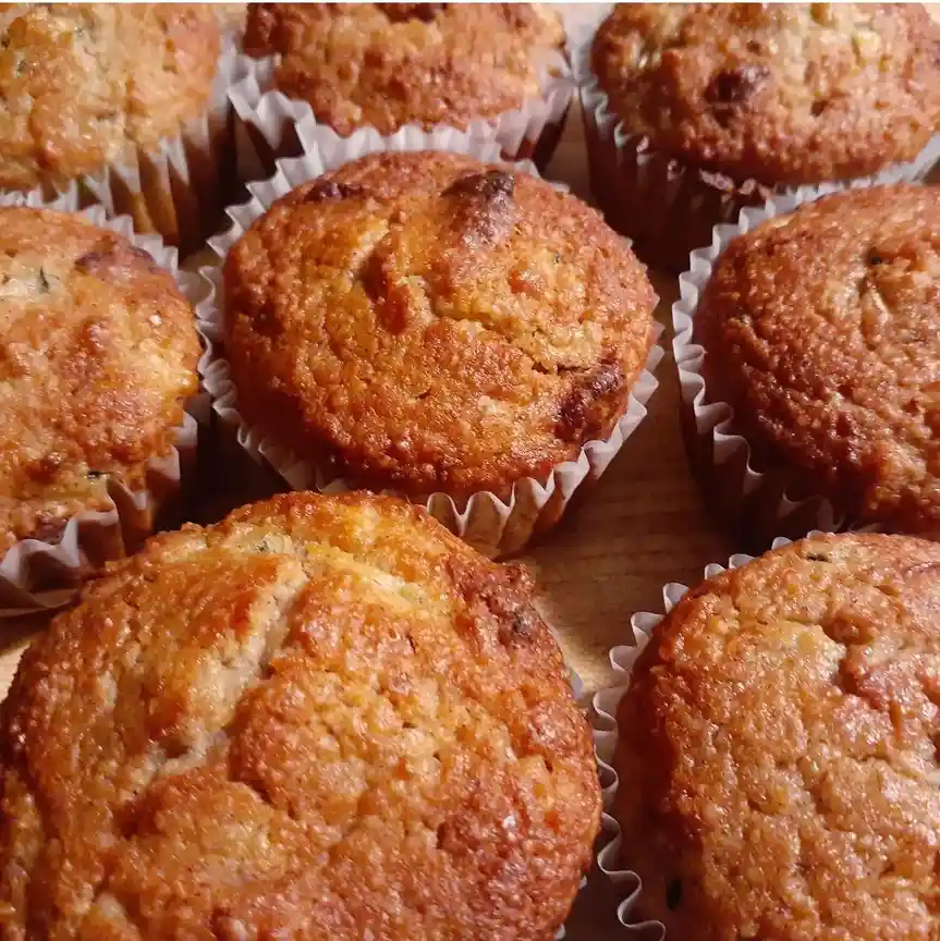 A close-up photo shows several golden-brown muffins in white paper liners, arranged on a wooden surface. The muffins have a…