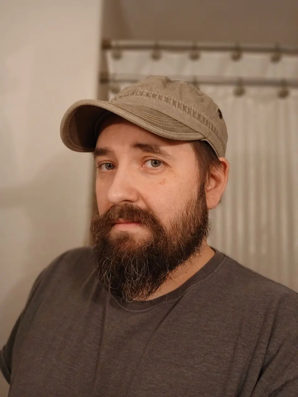 A man with a full beard and a khaki baseball cap looks directly at the camera with a neutral expression. He wears a dark…