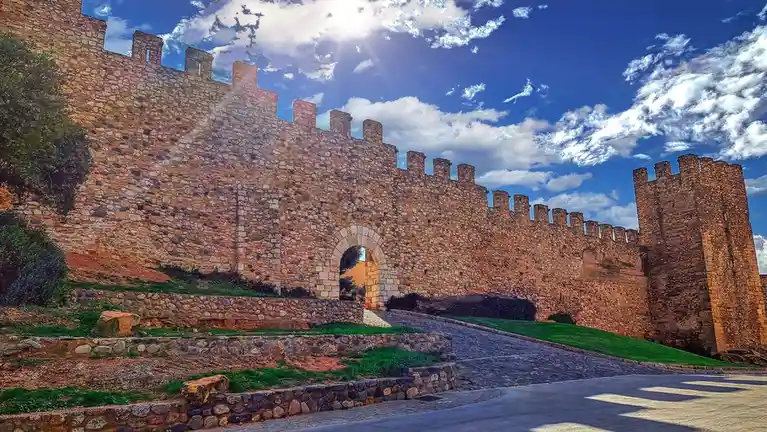 A wide-angle photo shows a large, ancient stone castle wall with crenellations under a bright blue sky with white clouds. A…