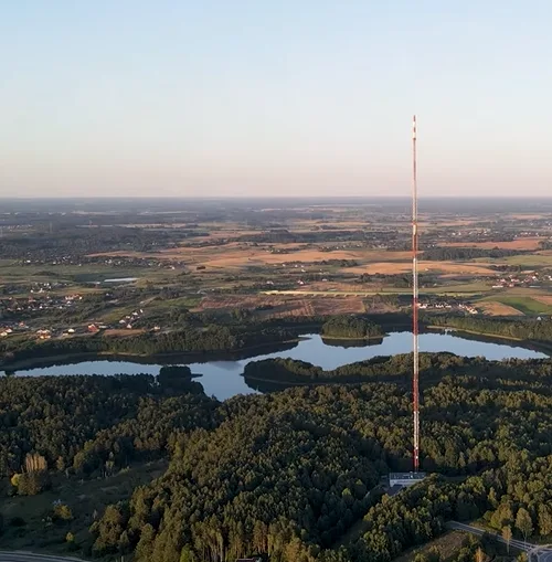 An aerial photo shows a tall, red-and-white striped communications tower standing prominently in a forested area next to a…