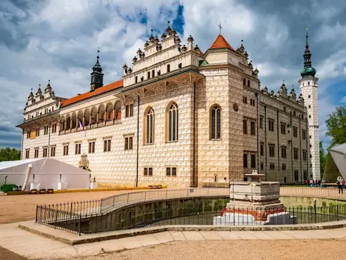 a grand, ornate stone castle with multiple towers and red-tiled roofs under a cloudy sky, featuring a small fountain in the…