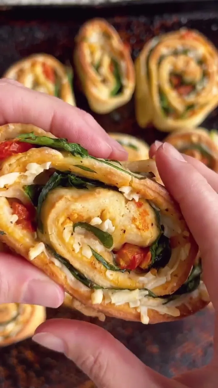 a close-up photo shows hands holding a baked, spiral-shaped food item with visible layers of cheese, spinach, and tomato…