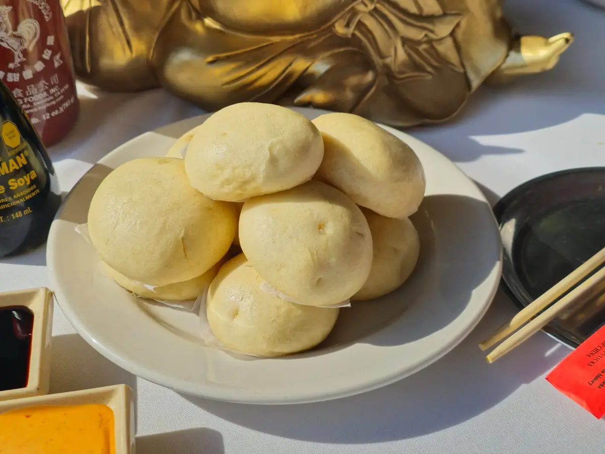 A plate of plain, steamed buns sits on a table next to soy sauce, chili sauce, chopsticks, and a golden Buddha statue. The…