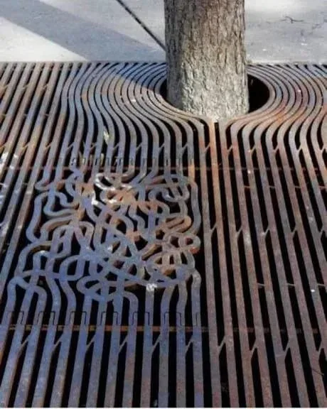 A close-up photo shows a tree trunk growing through a metal grate on pavement. The grate has straight, parallel bars on…