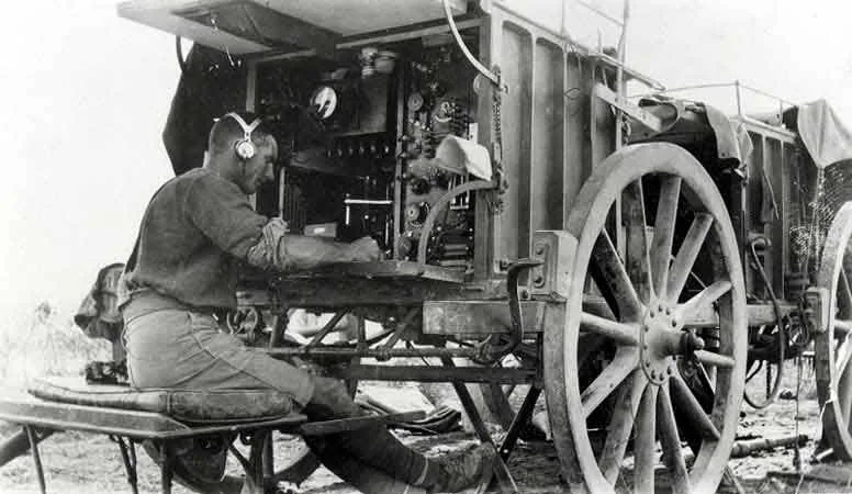 A black-and-white historical photo shows a man in early 20th-century military-style clothing sitting on a bench, wearing…