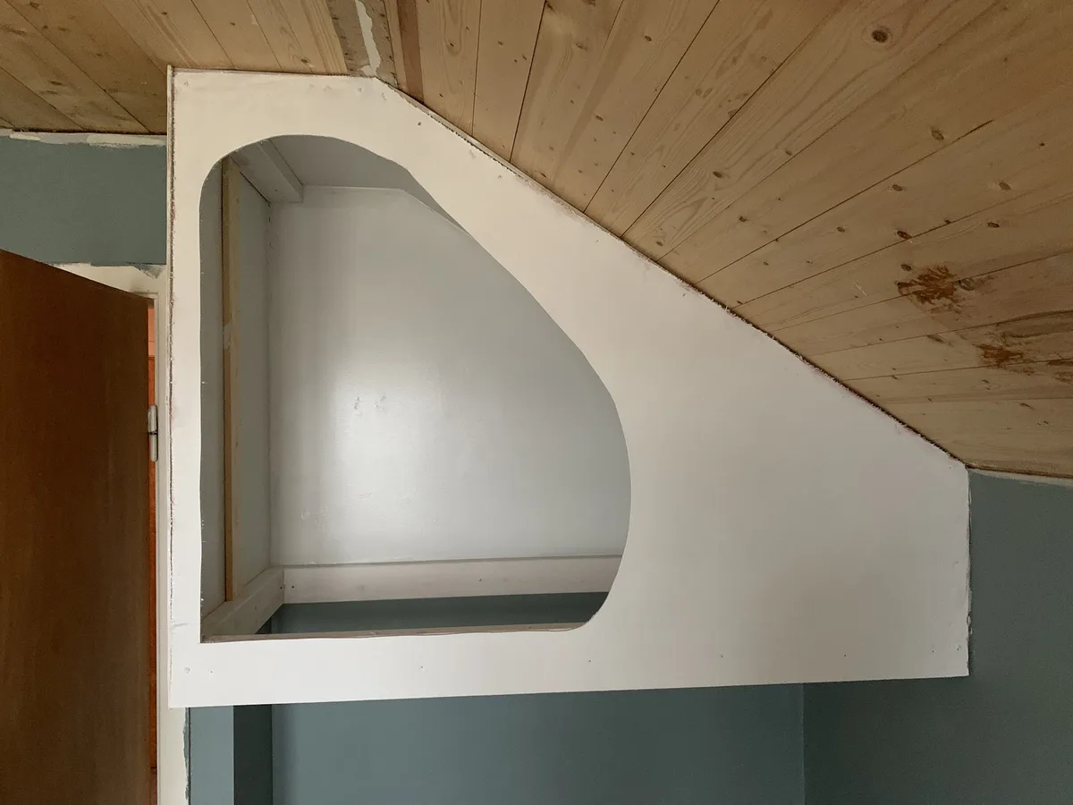 A photo shows a white, arched cutout in a wall, resembling a doorway but clearly not functional, set against a wooden…