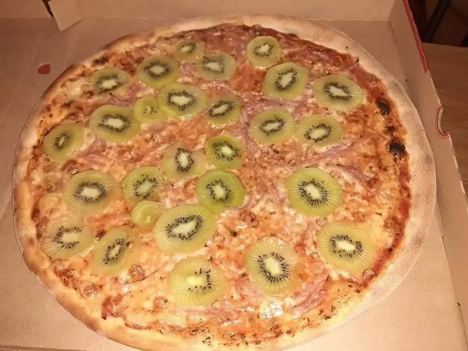 A pizza in a cardboard box is topped with numerous slices of kiwi fruit over cheese and ham, creating an unusual and…