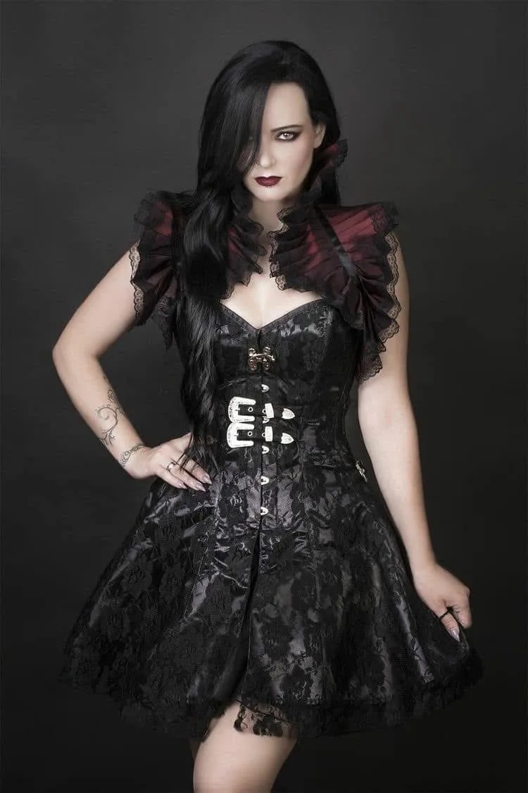 A woman with long black hair and dark makeup poses in a black lace gothic dress with a corset and ruffled collar, one hand…