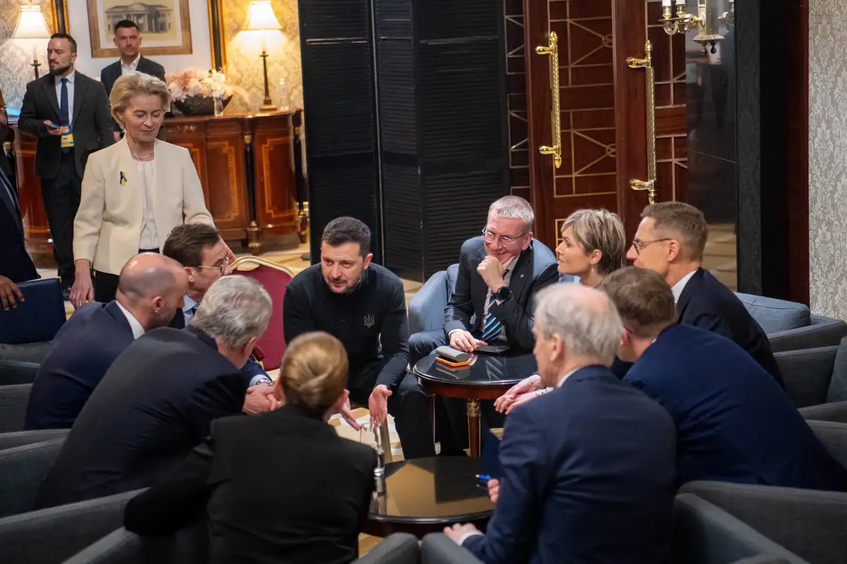 A group of formally dressed men and women are gathered around a small table in an ornate room, leaning in closely as if…