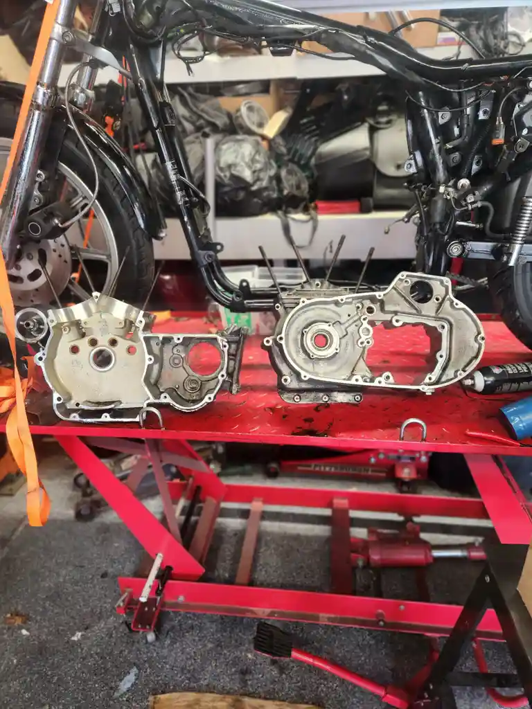 Two disassembled motorcycle engine cases sit side-by-side on a red workbench in a garage, one appearing heavily worn and the…