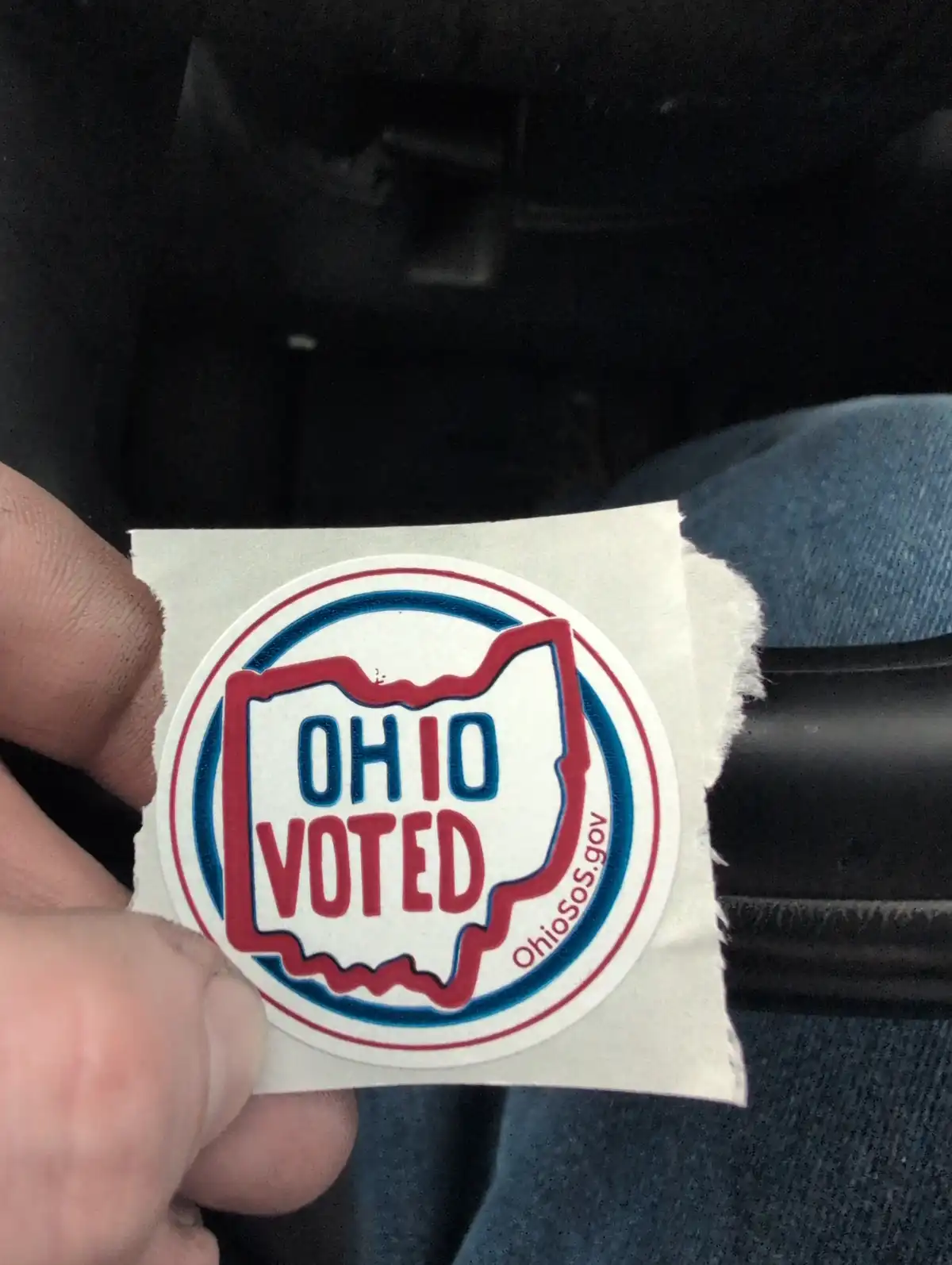 A hand holds a circular 'OHIO VOTED' sticker featuring the state outline in red and blue, with 'Ohiosos.gov' along the…