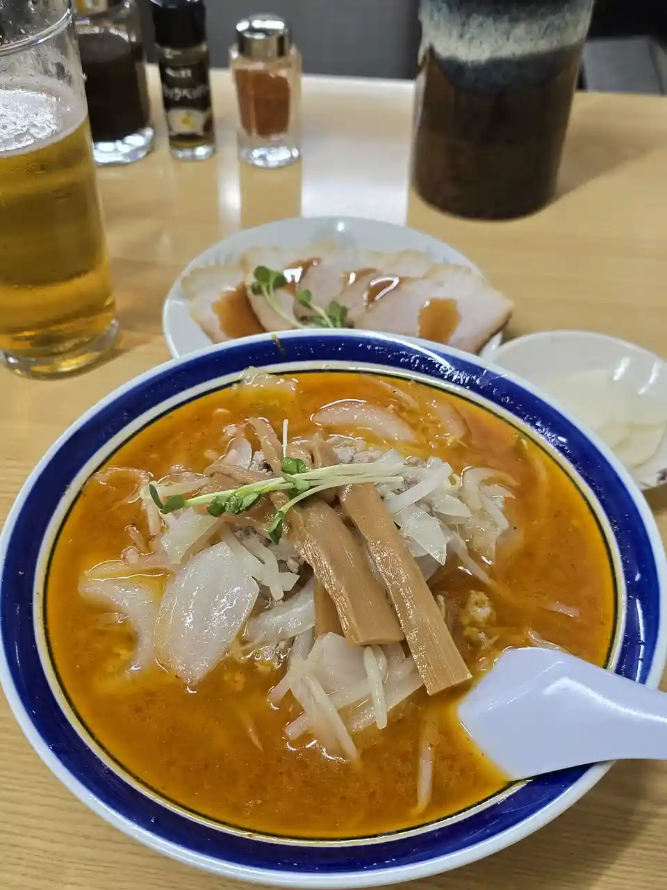 A close-up photo shows a steaming bowl of ramen with a rich orange broth, topped with bean sprouts, bamboo shoots, and…