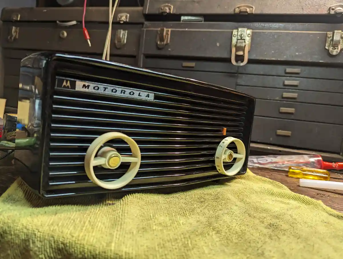 A vintage black Motorola radio with cream-colored tuning knobs sits on a green cloth in a workshop, surrounded by toolboxes…