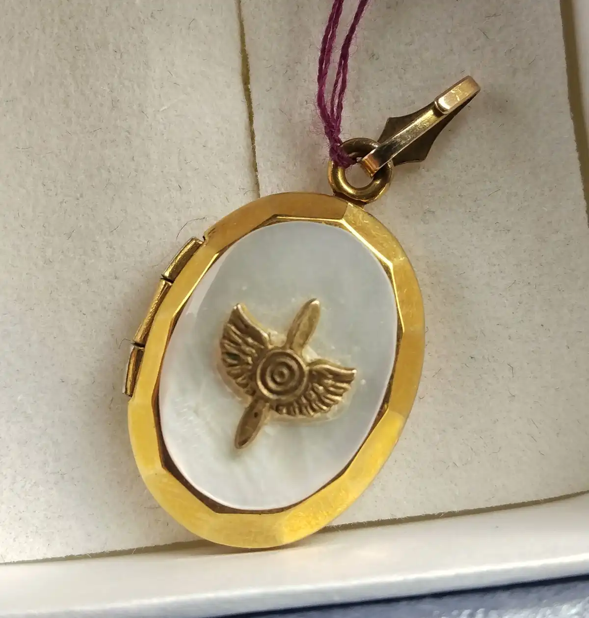 A close-up photo shows an oval gold locket with a white mother-of-pearl interior, featuring a small gold emblem of a winged…