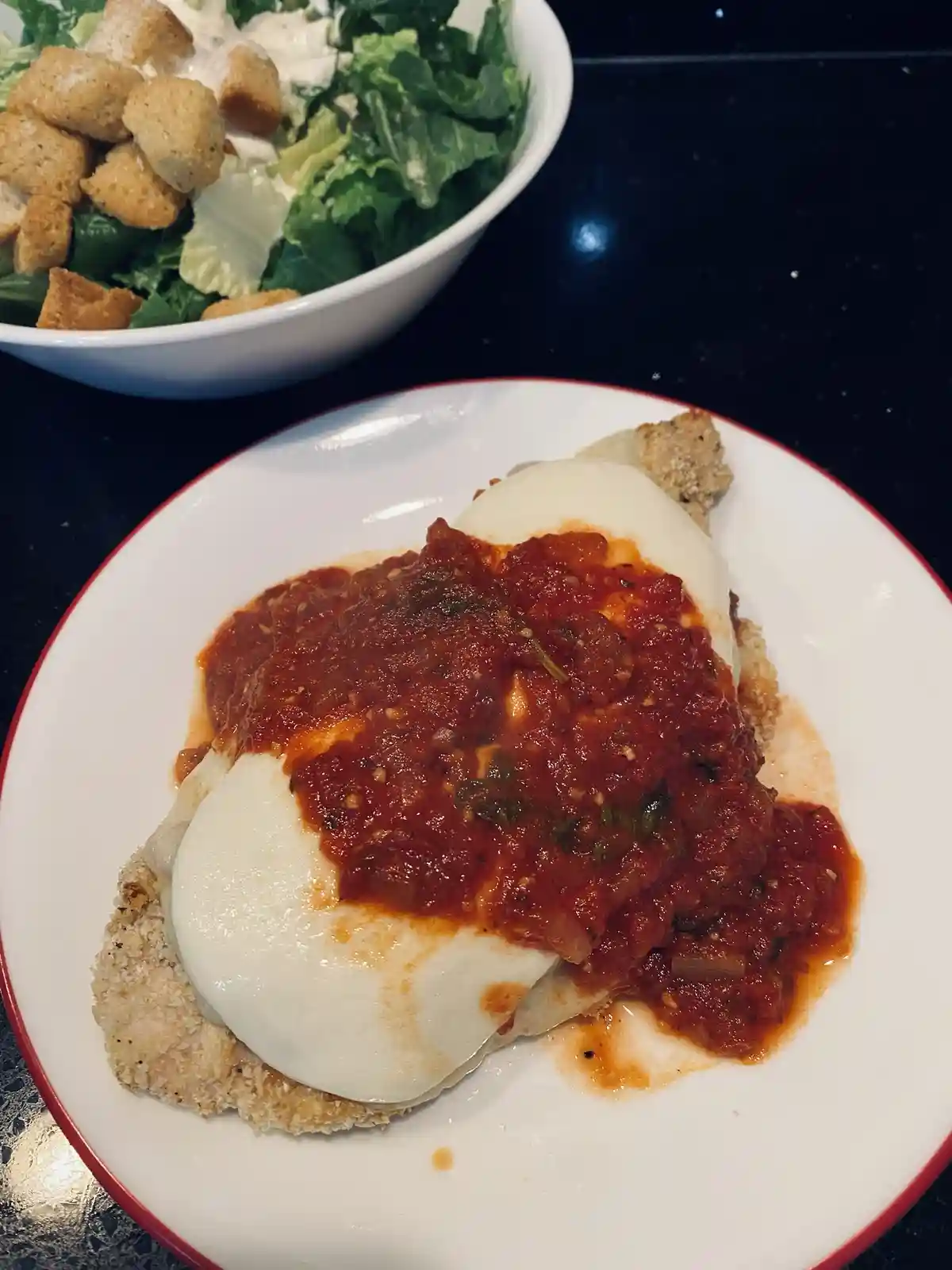 A close-up photo shows a plate of breaded chicken topped with tomato sauce and melted mozzarella cheese, next to a bowl of…