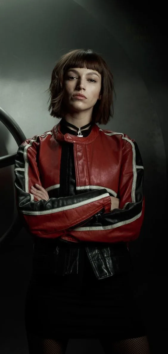 A woman with short brown hair and bangs stands with her arms crossed, wearing a red and black leather jacket with white…