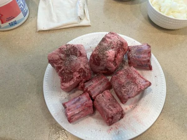 A white plate holds several raw, bone-in meat pieces, likely ribs or short ribs, generously seasoned with black pepper. The…