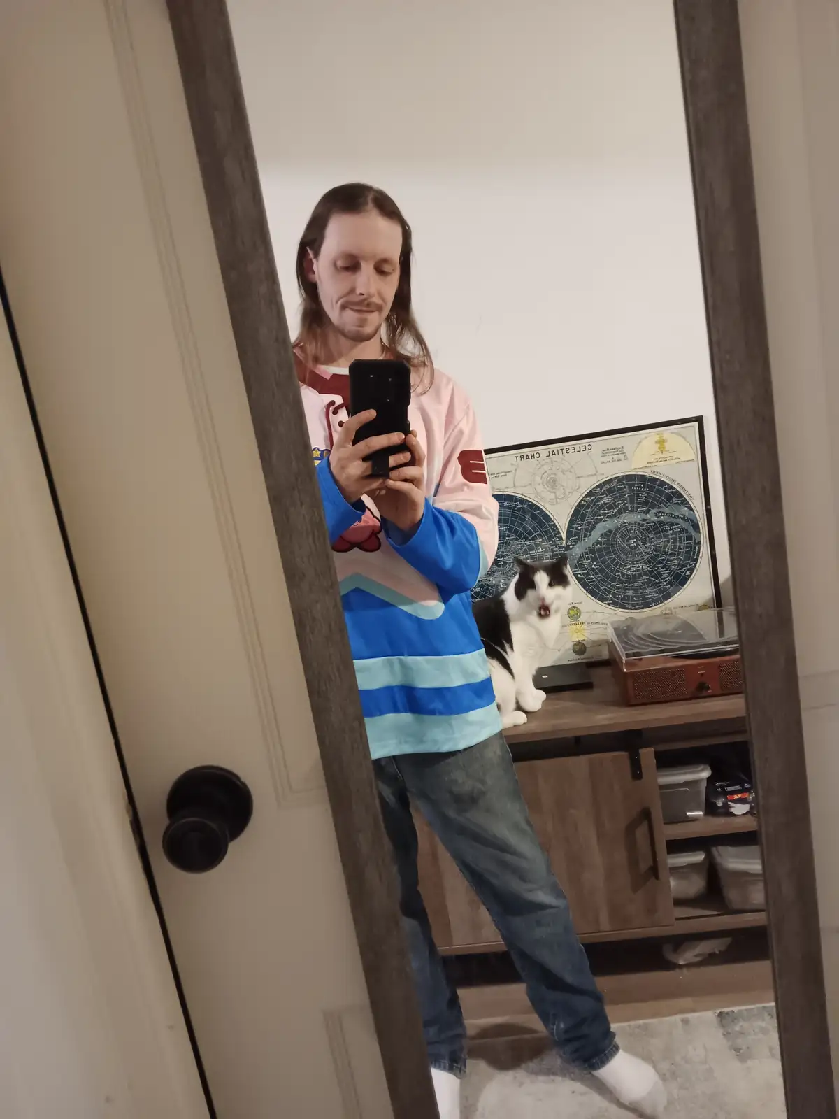 A person with long hair takes a mirror selfie while wearing a colorful, layered shirt and jeans, holding a phone. Behind…