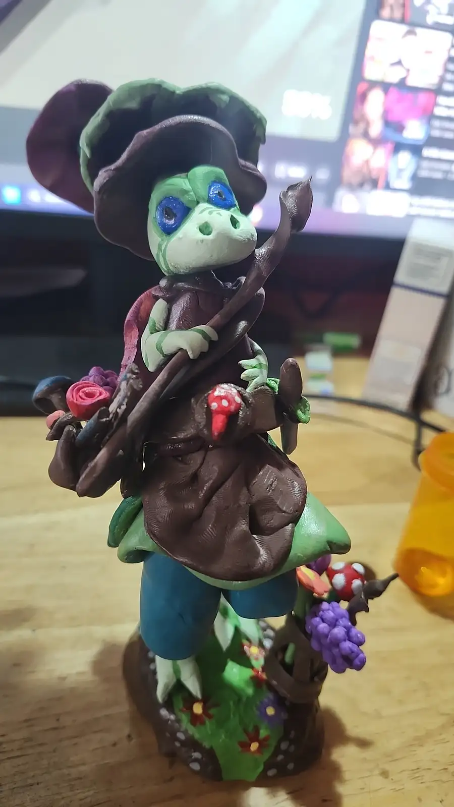 A handmade clay figurine of a green frog wearing a large brown witch's hat and cloak, holding a staff and surrounded by…