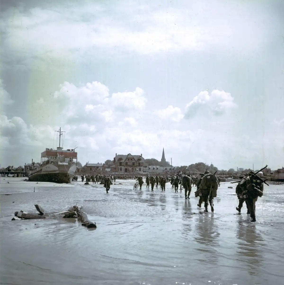 Soldiers in WWII-era uniforms wade through shallow water on a beach, carrying gear and weapons, with a ship and coastal…