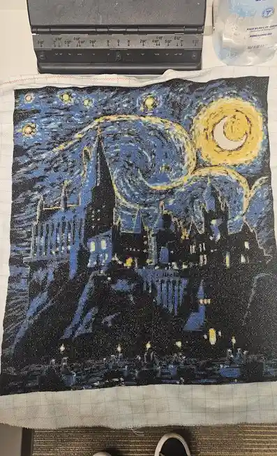 A completed needlepoint artwork depicting Vincent van Gogh's 'Starry Night' is laid flat on a table, showing the swirling…