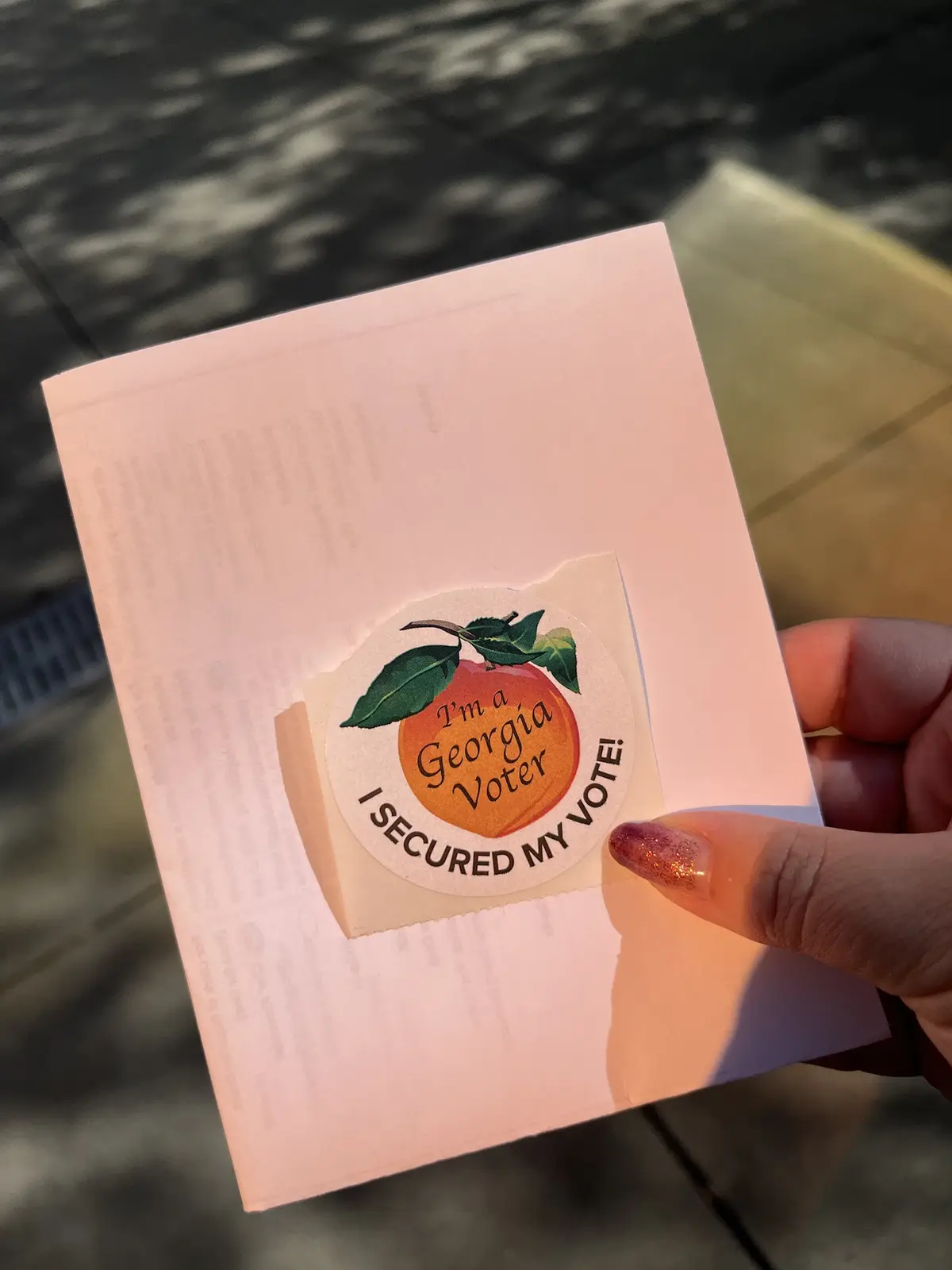 A hand holds a light pink paper envelope with a circular sticker on it. The sticker features a peach illustration with green…