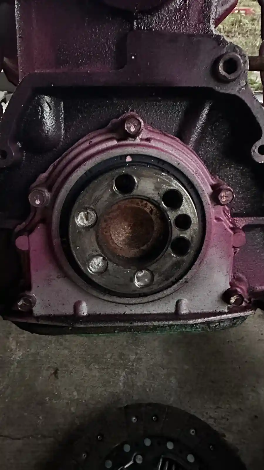 A close-up photo shows a dirty, rusty engine block with a pinkish-purple painted surface, featuring a central circular…