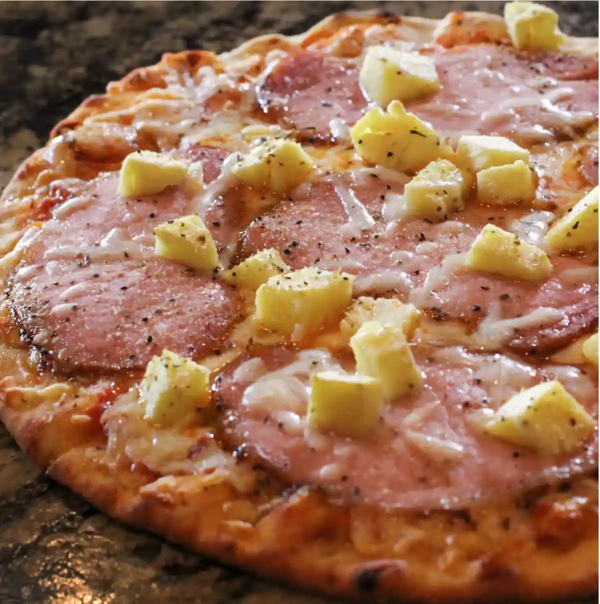A close-up photo of a freshly baked pizza with a golden-brown crust, topped with melted cheese, slices of ham, and chunks of…