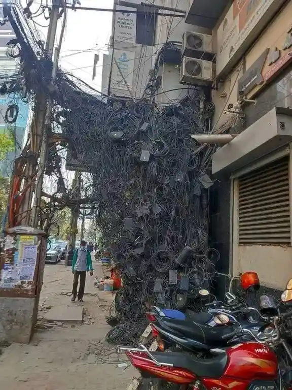 A chaotic tangle of black electrical wires, cables, and adapters cascades down a building wall and utility pole, covering…