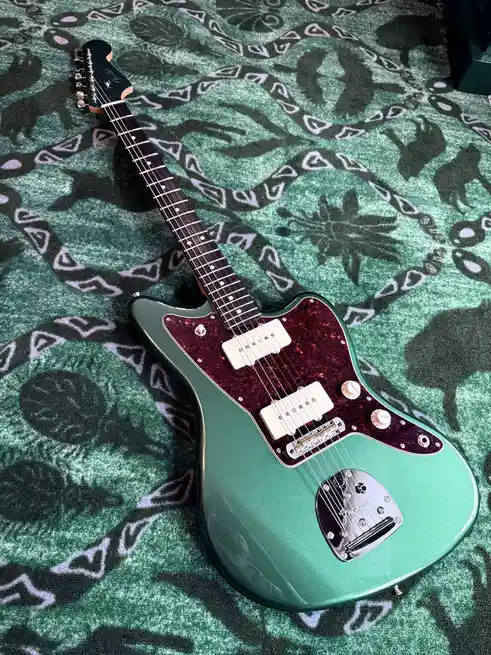 A green Fender electric guitar with a red pickguard rests on a green blanket featuring black and white tribal-style…