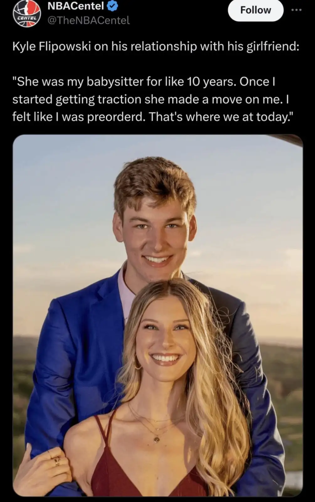 A photo of a smiling young man in a blue suit standing behind a smiling young woman in a red top, both looking at the…
