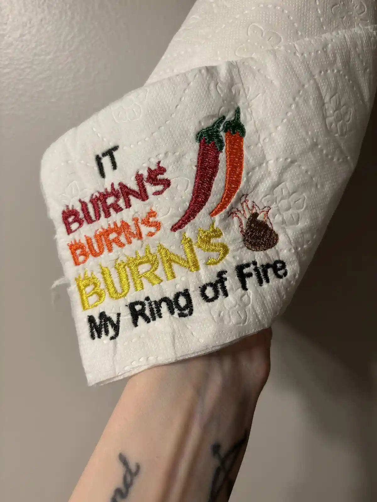 A close-up of a white paper towel roll held in a hand, featuring an embroidered design with three chili peppers and a poop…