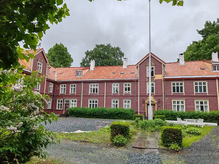 A red wooden building with orange tiled roofs and white-framed windows is shown under an overcast sky, with a flagpole in…