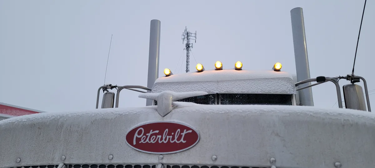 A close-up of the front of a white Peterbilt semi-truck, heavily covered in snow, with four glowing amber roof lights and…