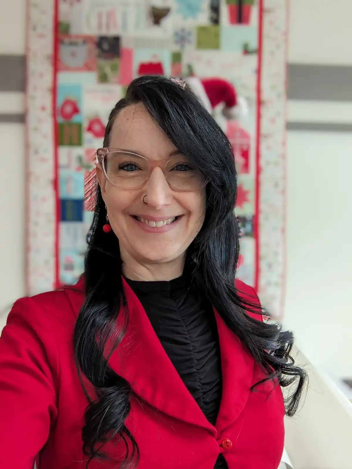 A woman with dark hair, wearing a red coat and glasses, smiles at the camera. Behind her is a colorful quilt with…