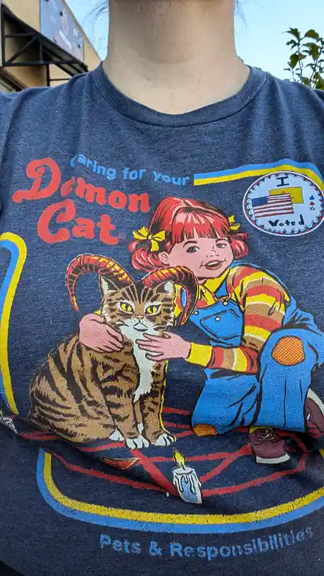 A person wears a blue t-shirt featuring a vintage-style illustration of a smiling girl in overalls petting a cat with devil…