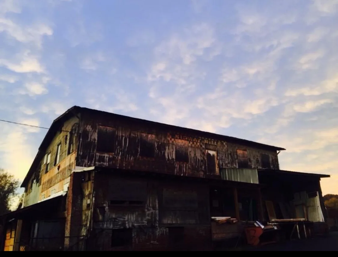 A low-angle photo of a dilapidated, rusted, two-story industrial building with boarded-up windows and peeling paint, set…