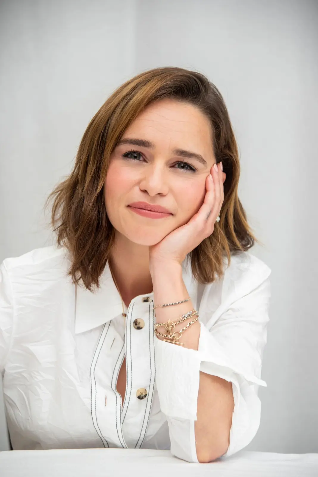 A woman with shoulder-length brown hair, wearing a white button-up shirt with black trim and multiple gold bracelets, rests…