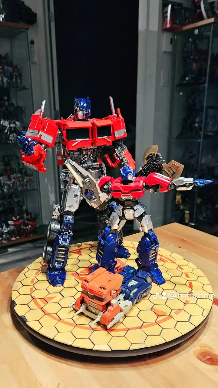 A large, detailed Optimus Prime action figure stands protectively over a smaller Optimus Prime figure, which is in turn…