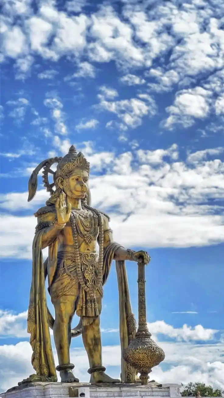 A large golden statue of Hanuman, the Hindu monkey god, stands against a bright blue sky with scattered white clouds. He is…