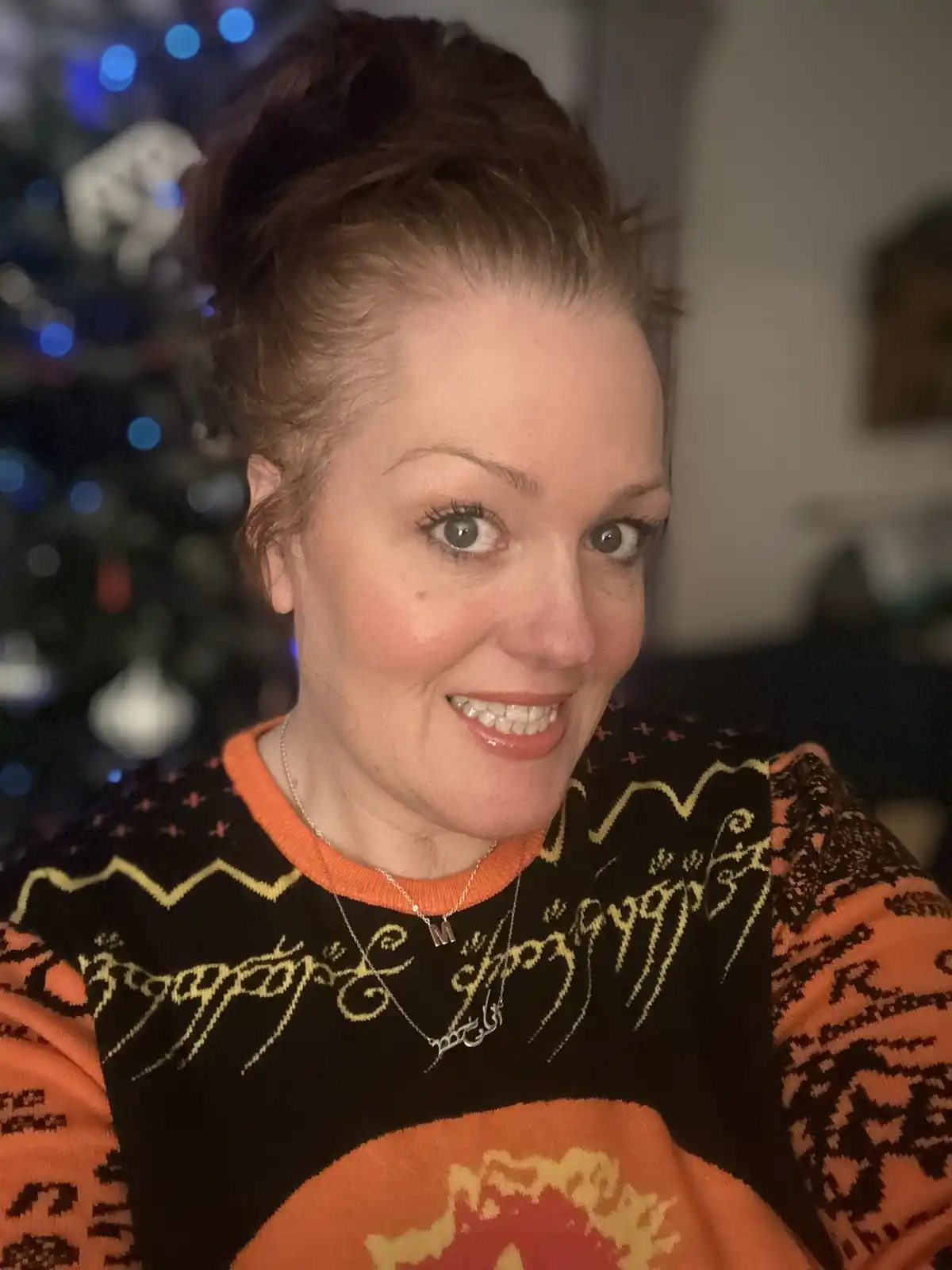 A woman with reddish hair in a bun smiles at the camera while wearing a black and orange holiday sweater with a large…