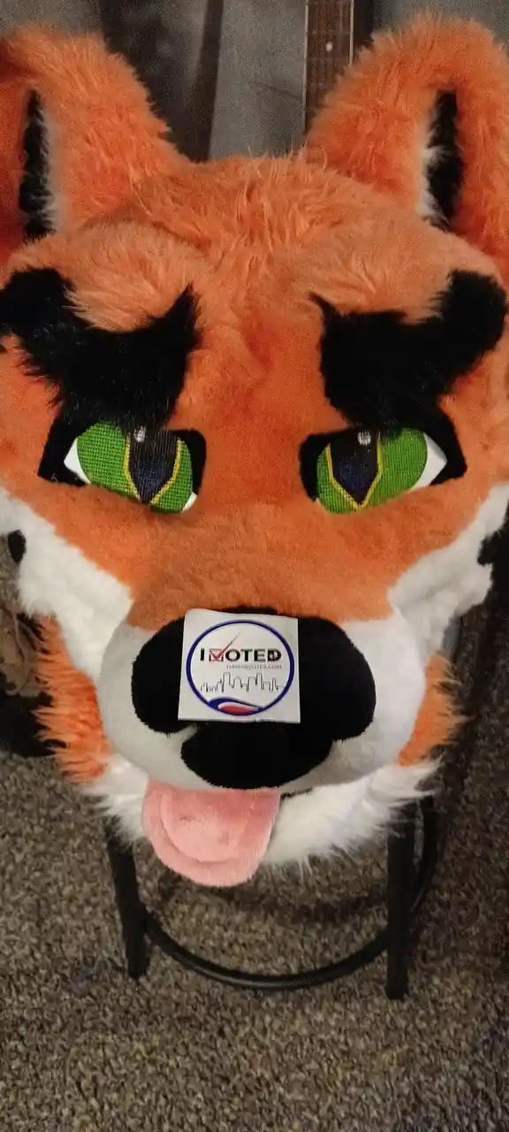 A close-up of a plush fox head costume with green eyes, black nose, and pink tongue sticking out, featuring an "I VOTED"…