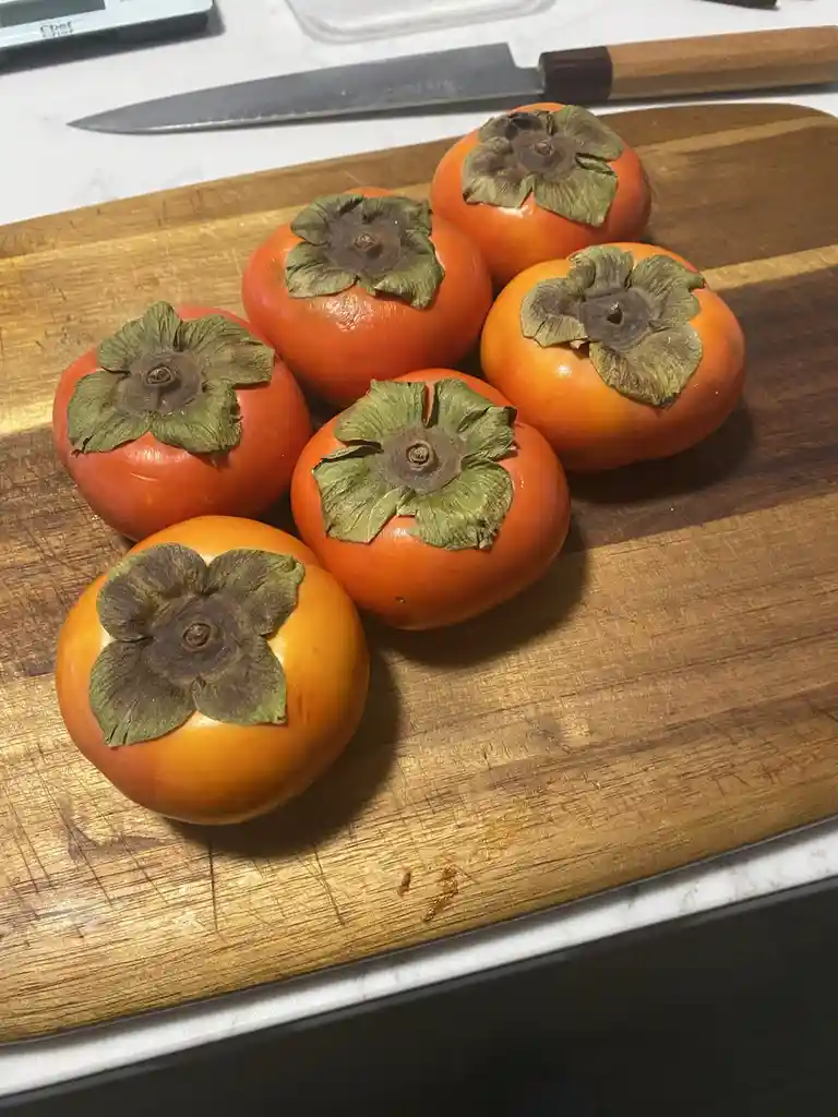Six ripe, orange persimmons with green leafy caps sit on a well-used wooden cutting board. A large kitchen knife rests in…