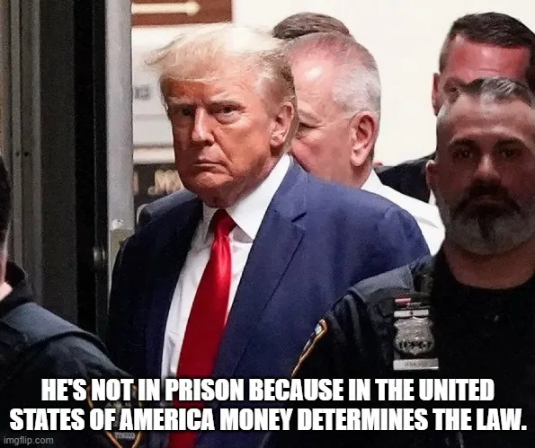 A photo of Donald Trump in a blue suit and red tie, looking stern while being escorted by security personnel. The overlaid…
